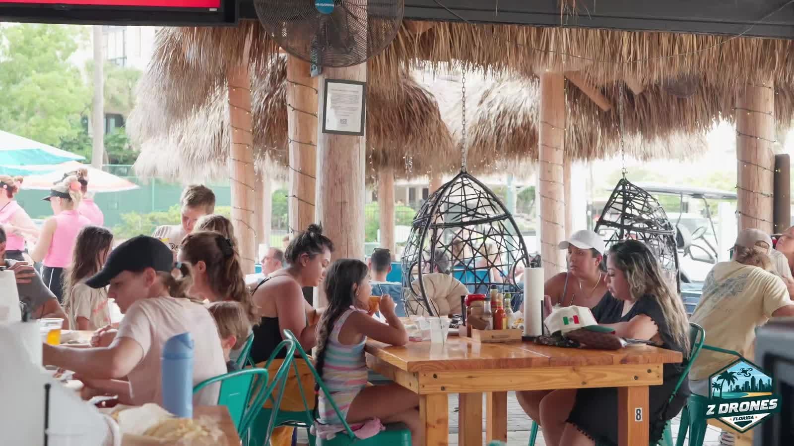 Busy covered terrace of a Gulf-front tiki lounge with guests seated at tables under a thatched roof