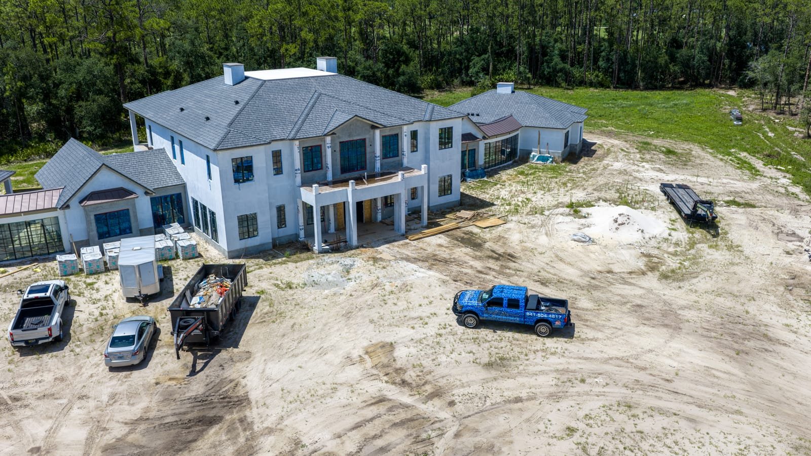Aerial view of a large two-storey custom home nearing completion on a cleared sand lot with contractor trucks on site
